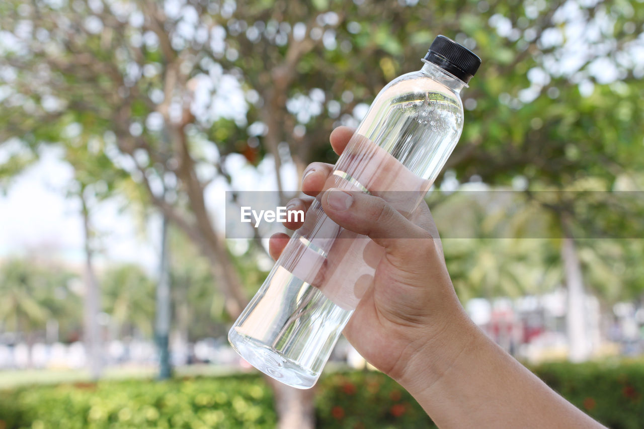 CLOSE-UP OF HAND HOLDING GLASS BOTTLE AGAINST BLURRED BACKGROUND