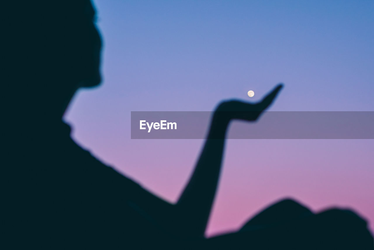 Close-up of silhouette person holding moon against clear blue sky at dusk