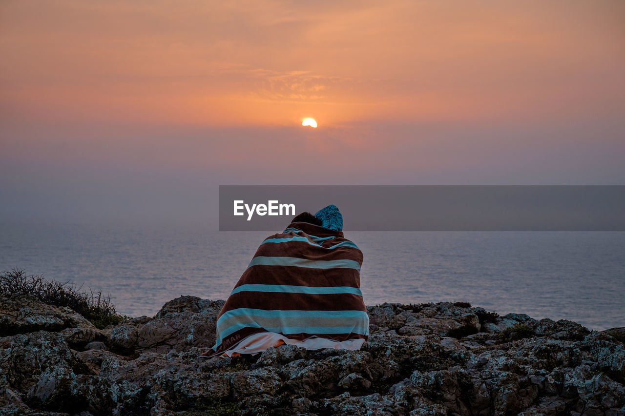 Friends sitting on rocks against sea during sunset