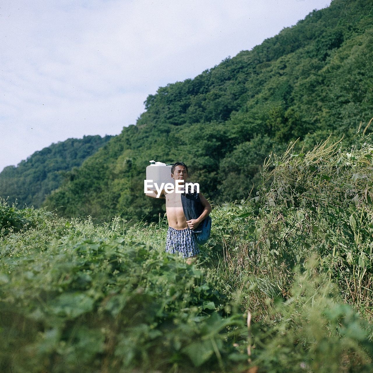 Shirtless young man carrying container while standing amidst plants against mountain
