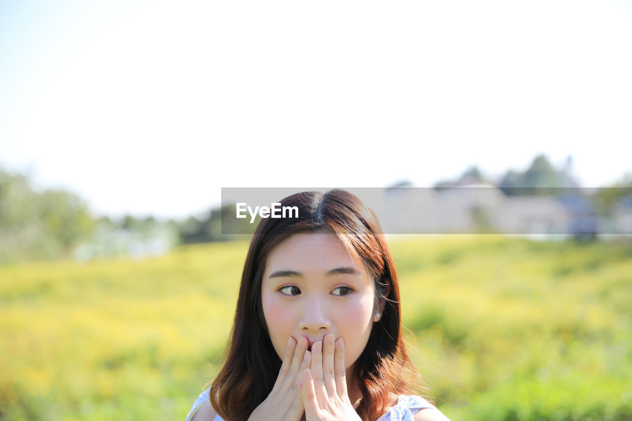 Surprised young woman covering mouth while looking away against sky