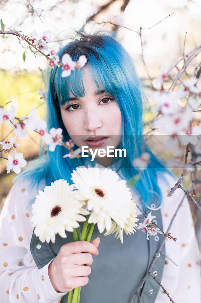 Modern trendy female with blue hair holding bouquet of fresh flowers and looking at camera while standing in blooming spring garden