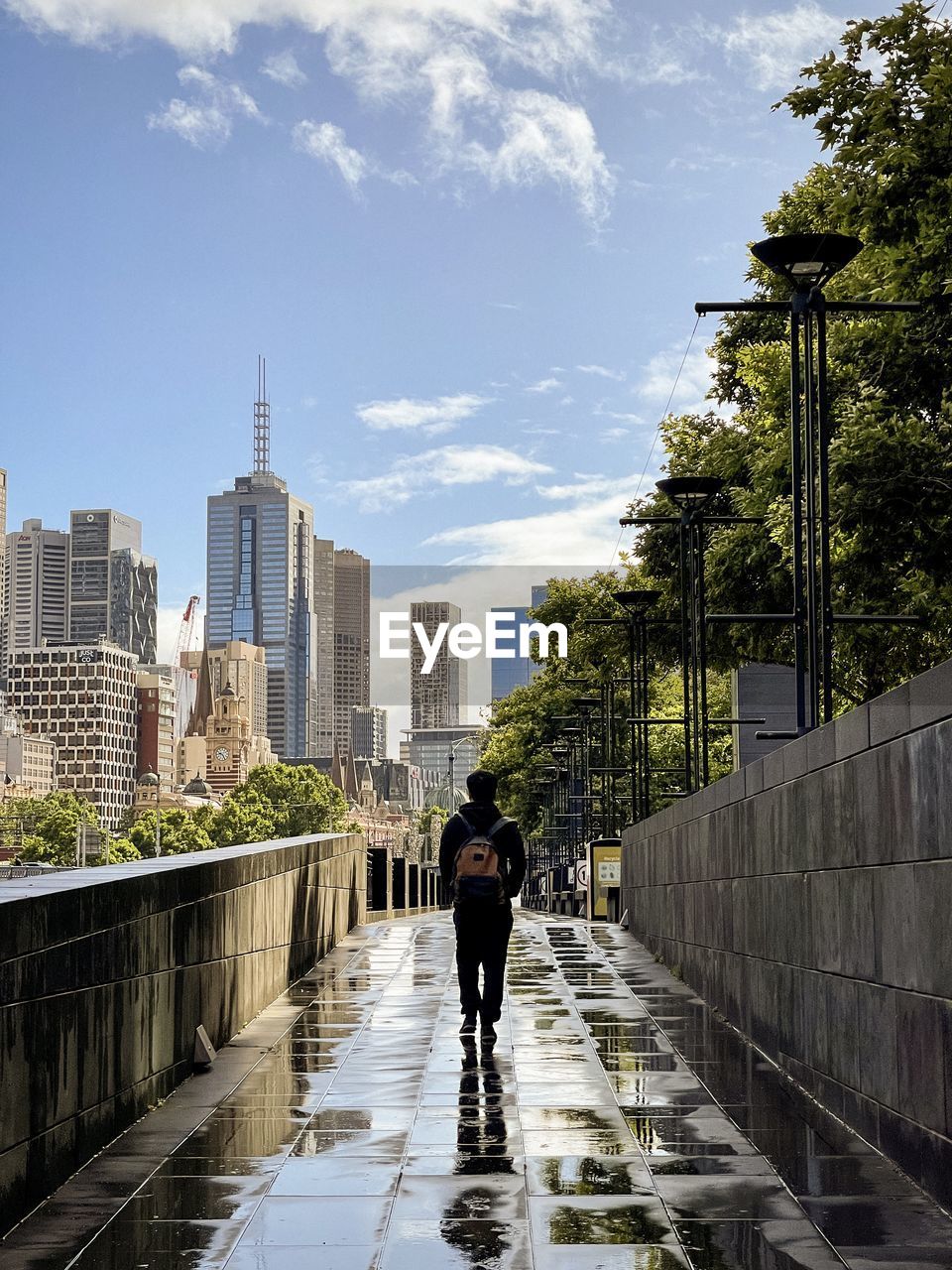 Rear view of man walking on paved footpath against buildings and urban skyline.