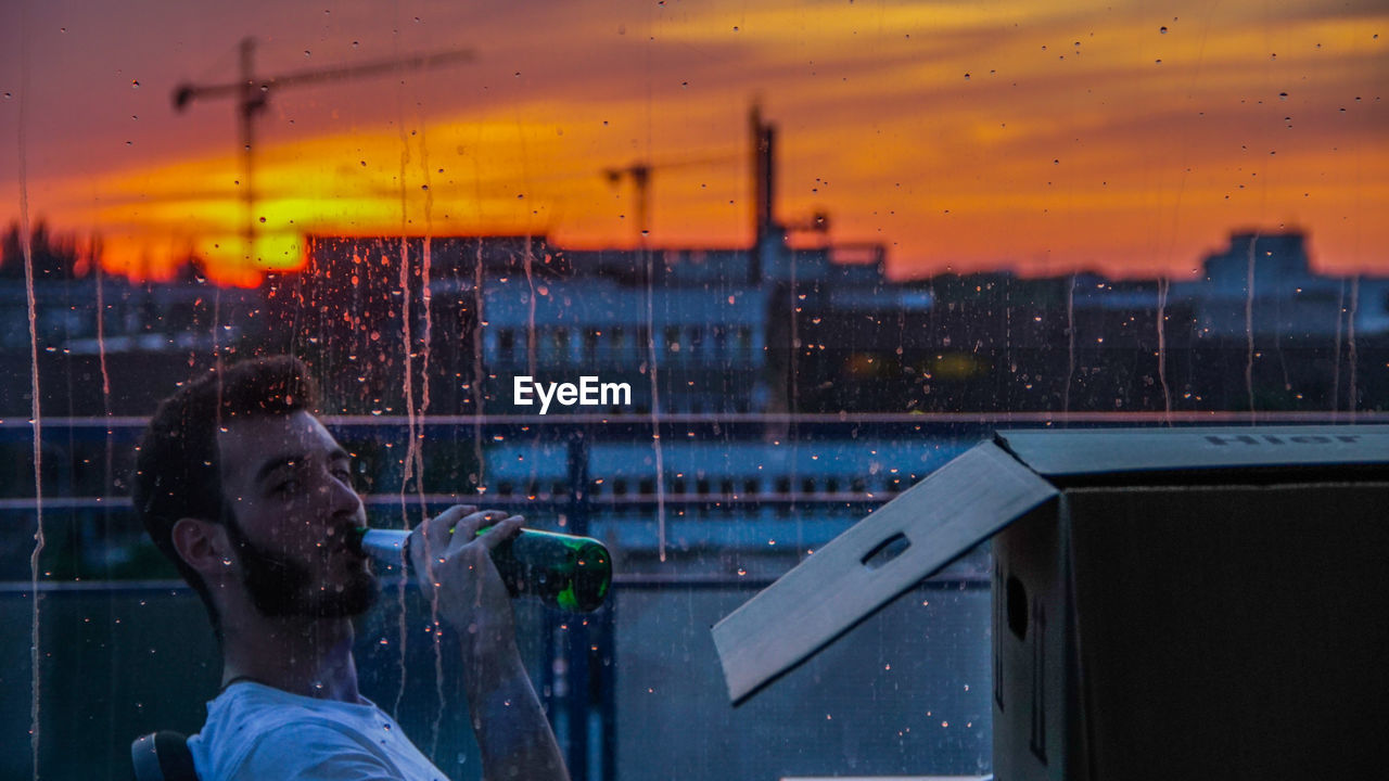 Young man drinking in balcony seen through wet window during sunset