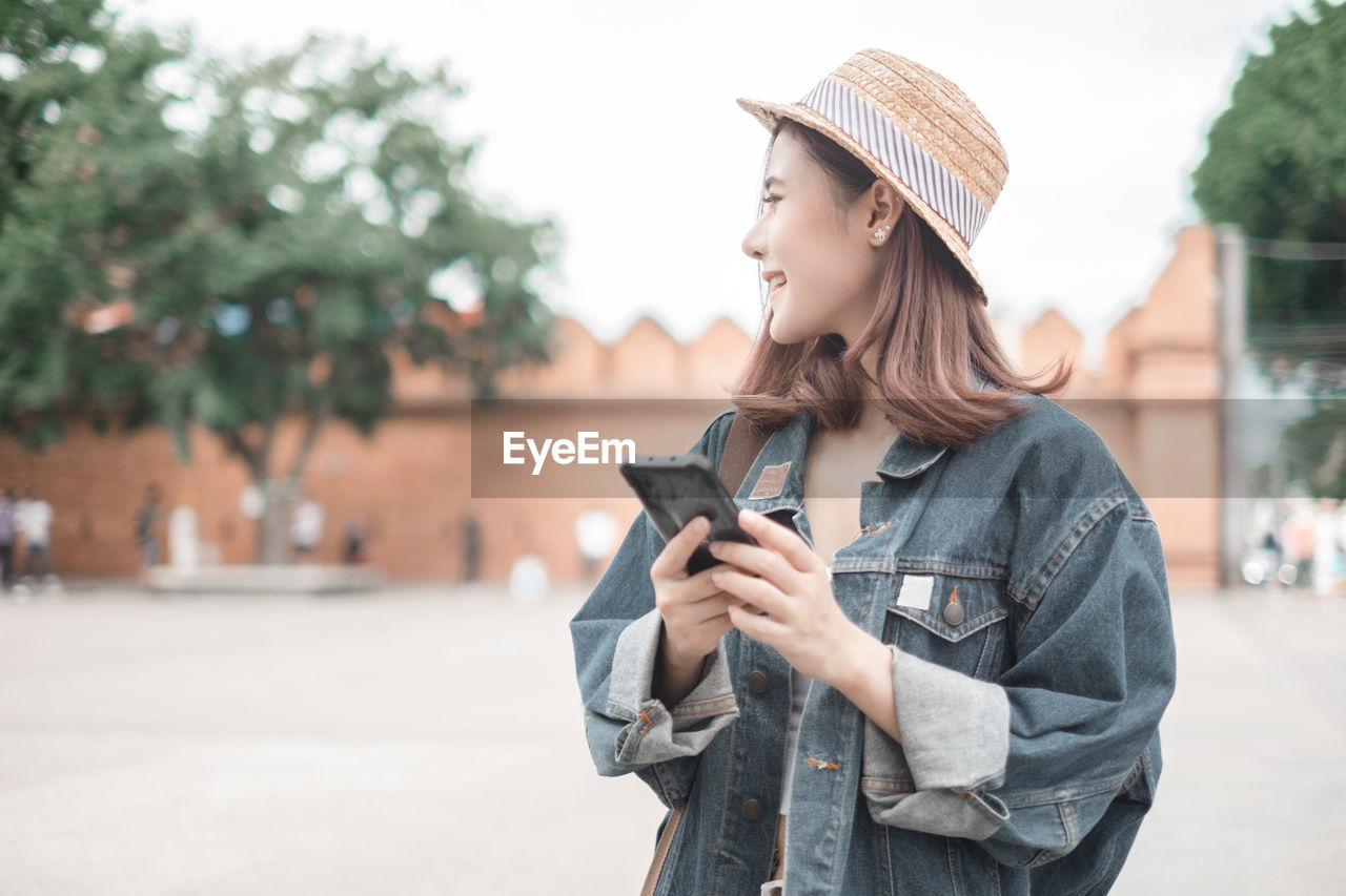 YOUNG WOMAN USING SMART PHONE WHILE STANDING ON CITY