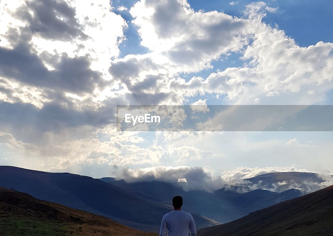 REAR VIEW OF MAN STANDING BY MOUNTAIN AGAINST SKY