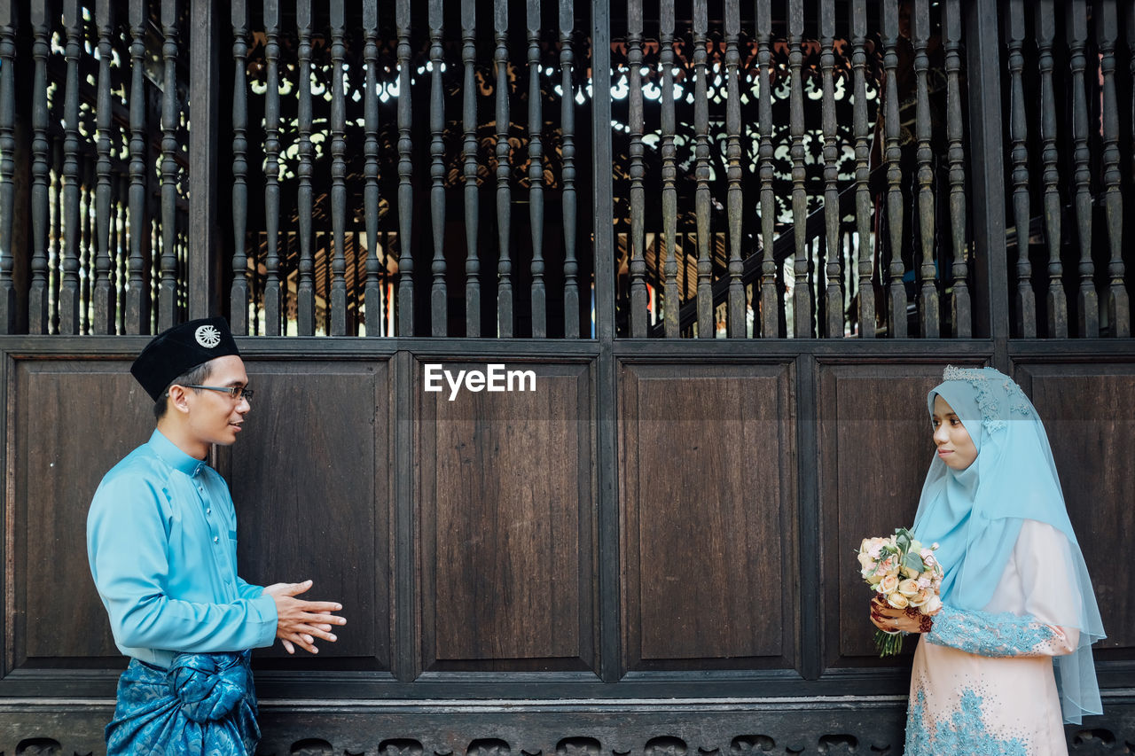 Wedding couple wearing traditional clothing while standing by wooden wall