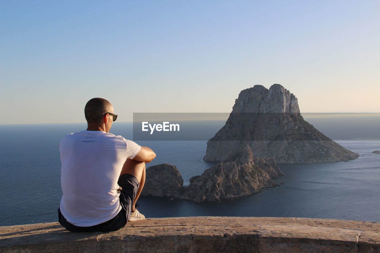 Rear view of young man sitting on retaining wall against seascape