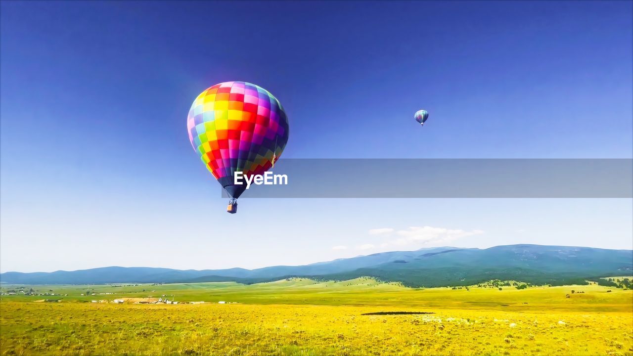hot air balloon, balloon, air vehicle, flying, mid-air, transportation, hot air ballooning, environment, aircraft, adventure, landscape, multi colored, sky, vehicle, mountain, nature, travel, blue, scenics - nature, beauty in nature, land, mode of transportation, tranquility, field, tranquil scene, mountain range, cloud, plant, rural scene, toy, journey, clear sky, non-urban scene, yellow, ballooning festival, taking off, travel destinations, outdoors, morning, day, leisure activity, vacation, no people, trip, motion, grass, holiday, sports, wind, basket, tourism, sunny, green, joy, summer, vibrant color, horizon over land