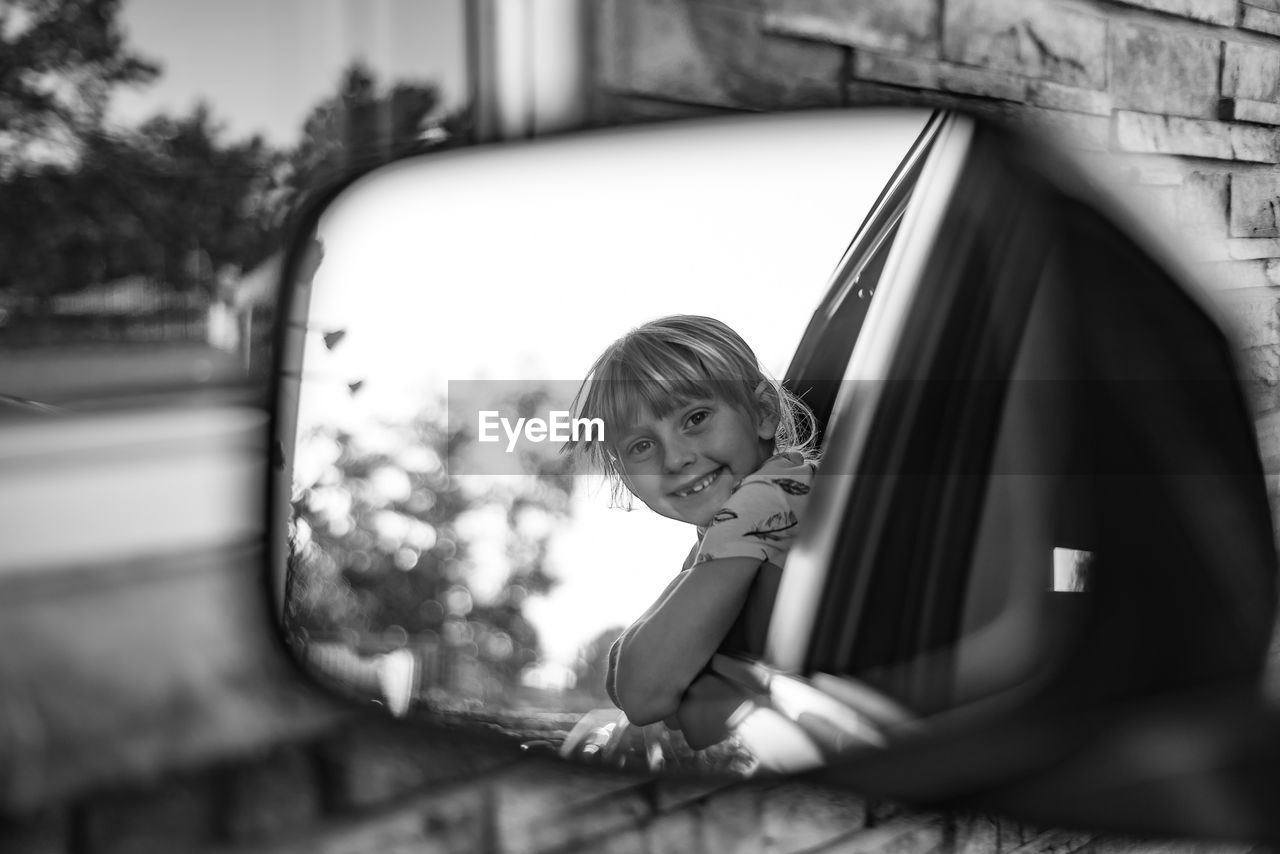 Portrait of cheerful girl leaning out from car window seen in side-view mirror
