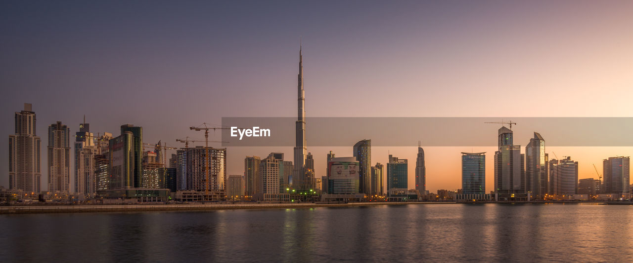Sea against burj khalifa amidst towers against sky at sunset