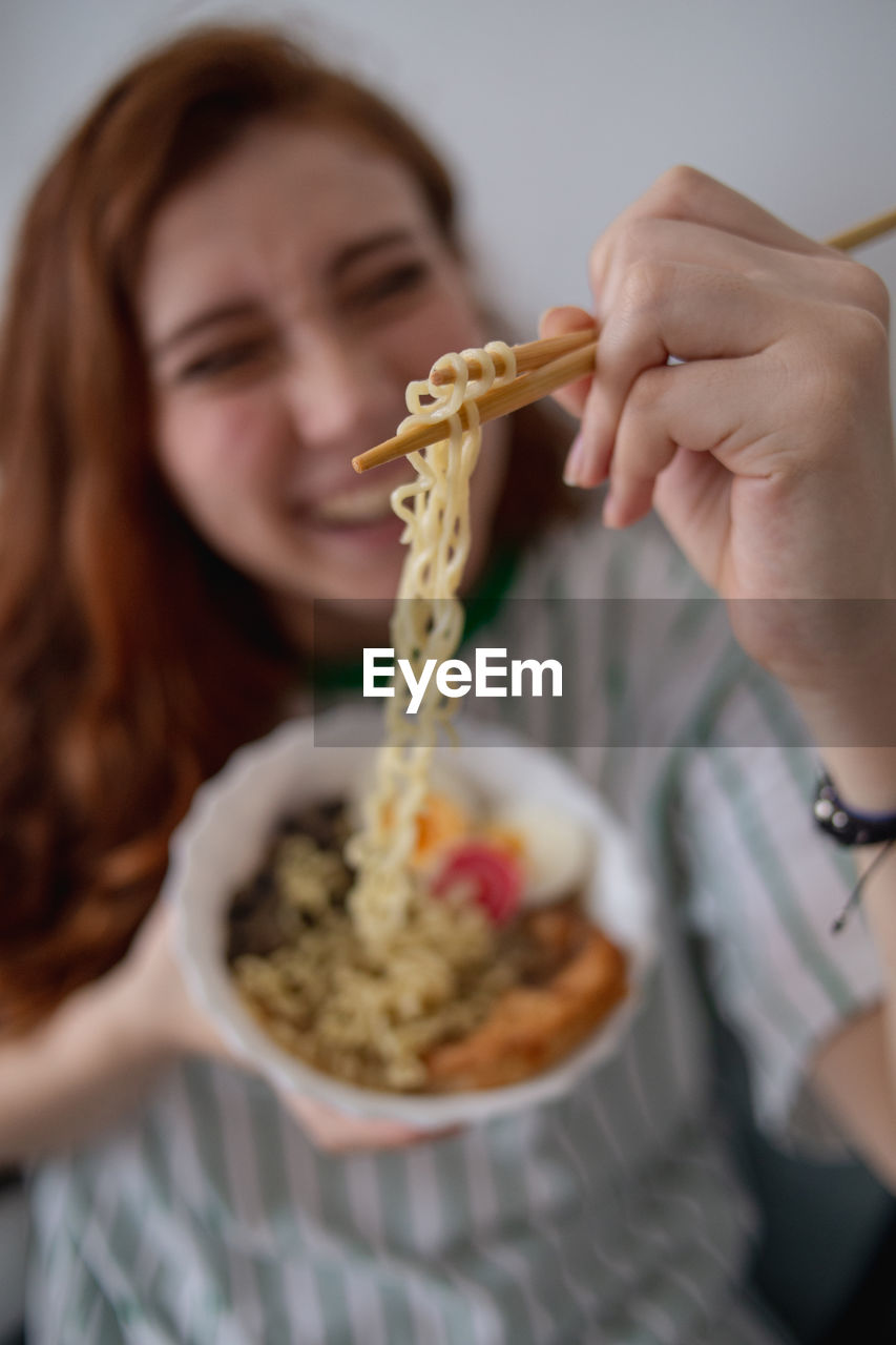 Blurred cheerful redhead female laughing and picking noodles from bowl of tasty ramen while sitting on couch at home