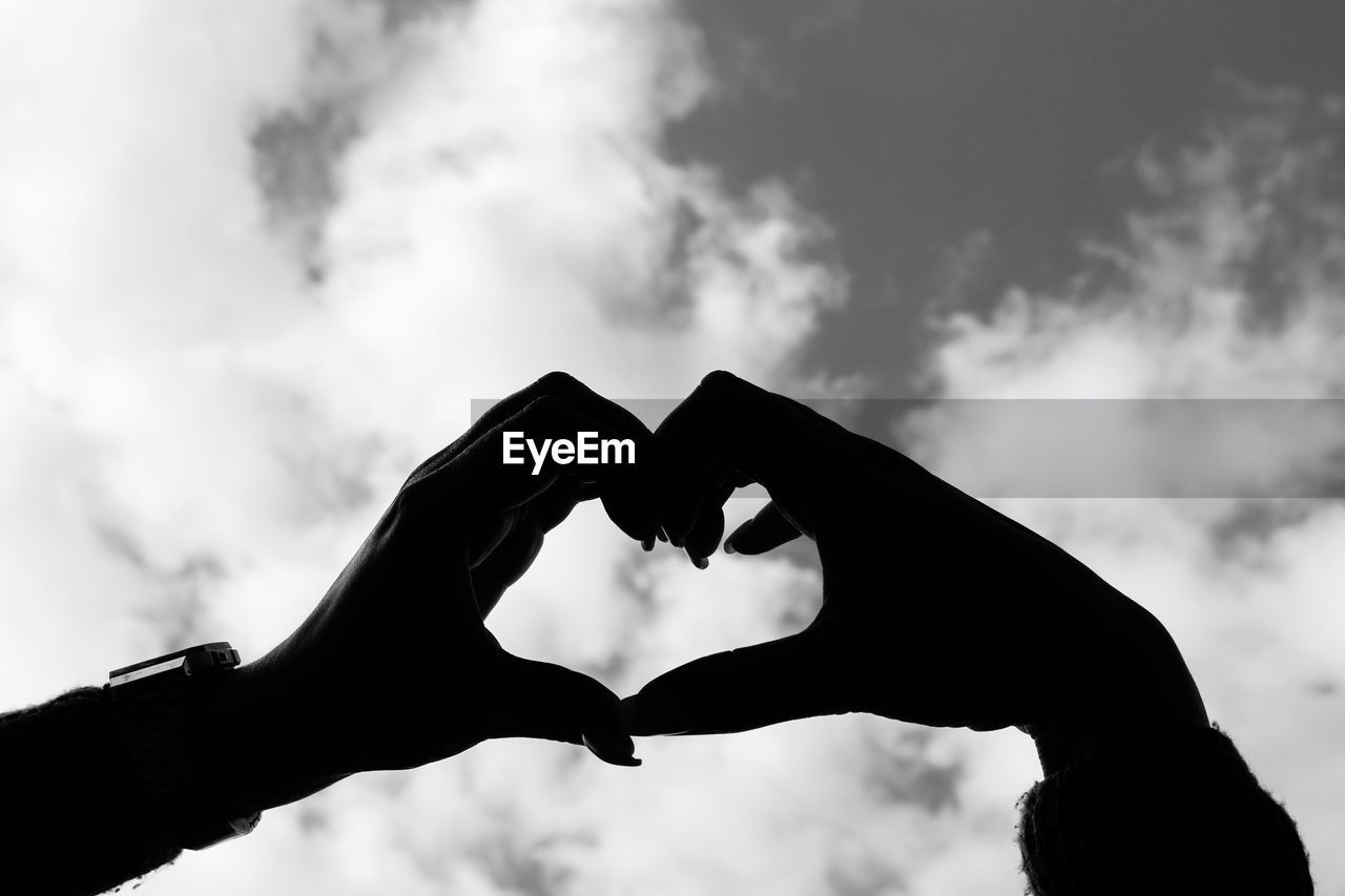 Cropped hands of woman making heart shape against cloudy sky