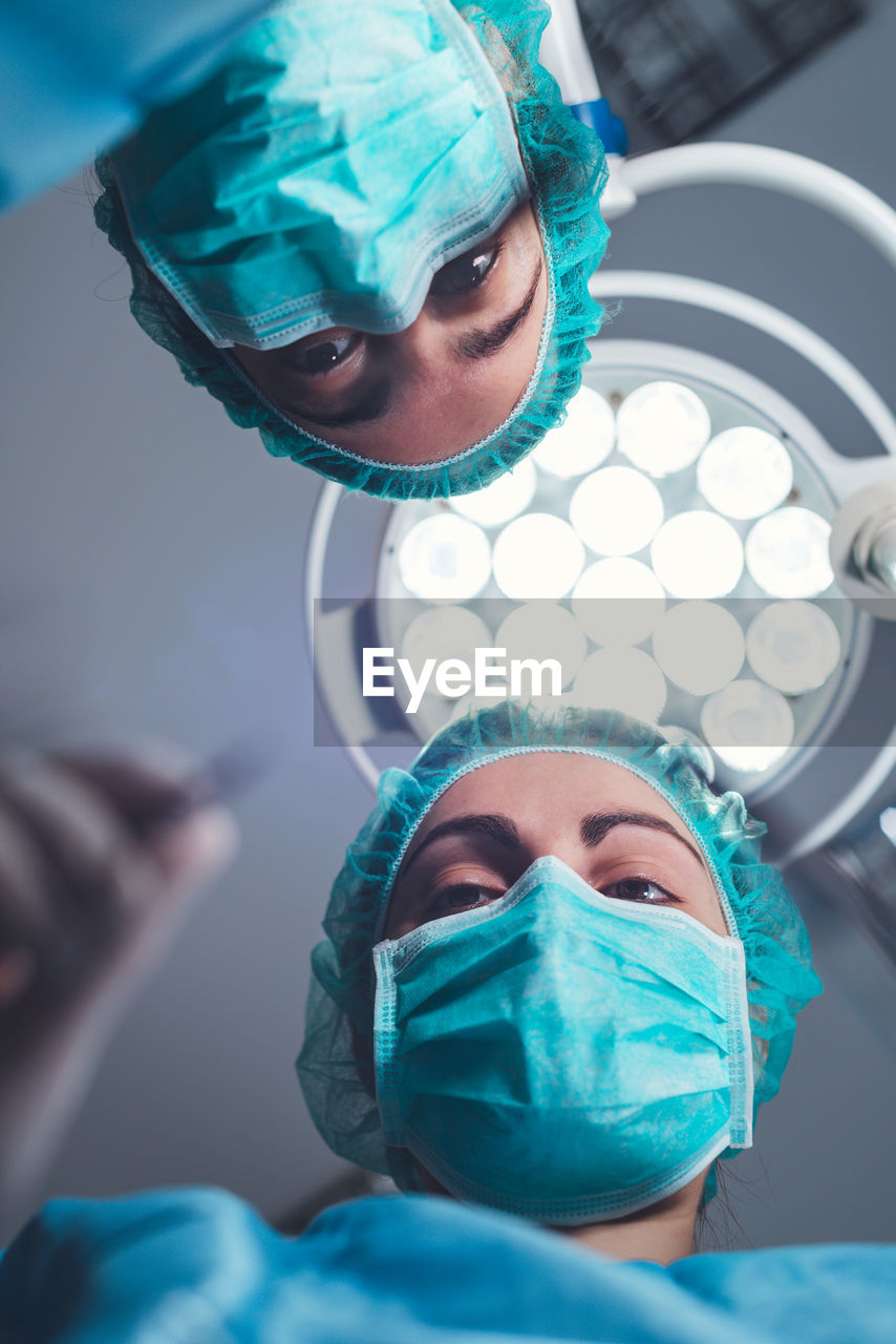 From below female surgeons in medical uniform using professional tools while standing under bright light in operating theater