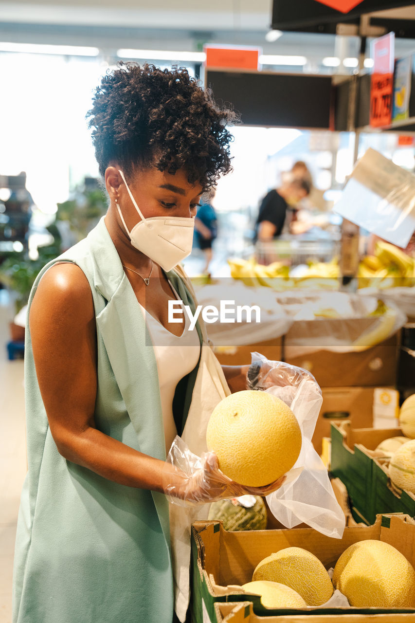 Side view of african american female customer in protective mask and gloves picking ripe watermelon from box while making purchases in supermarket during coronavirus pandemic