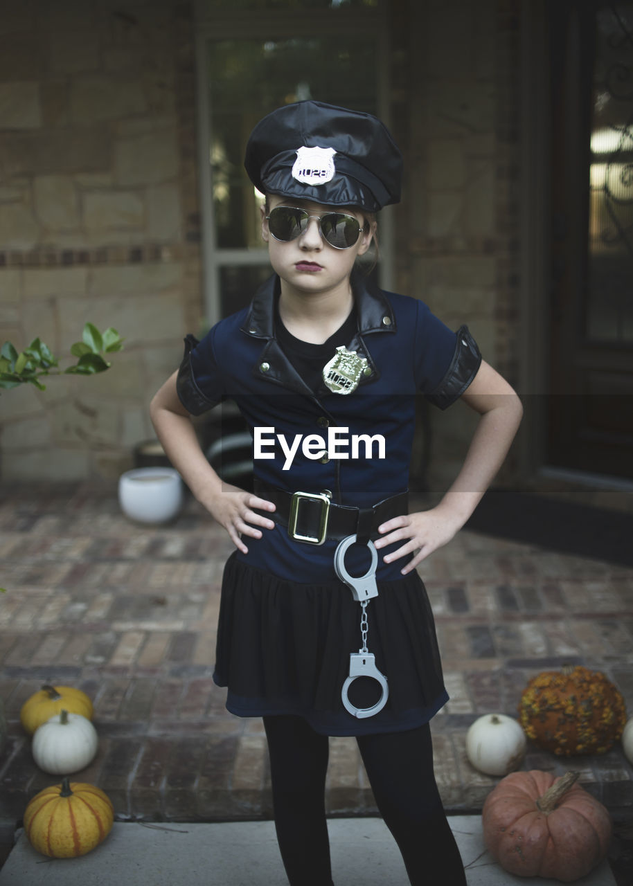 Portrait of confident girl in police costume with hands on hip standing against house during halloween