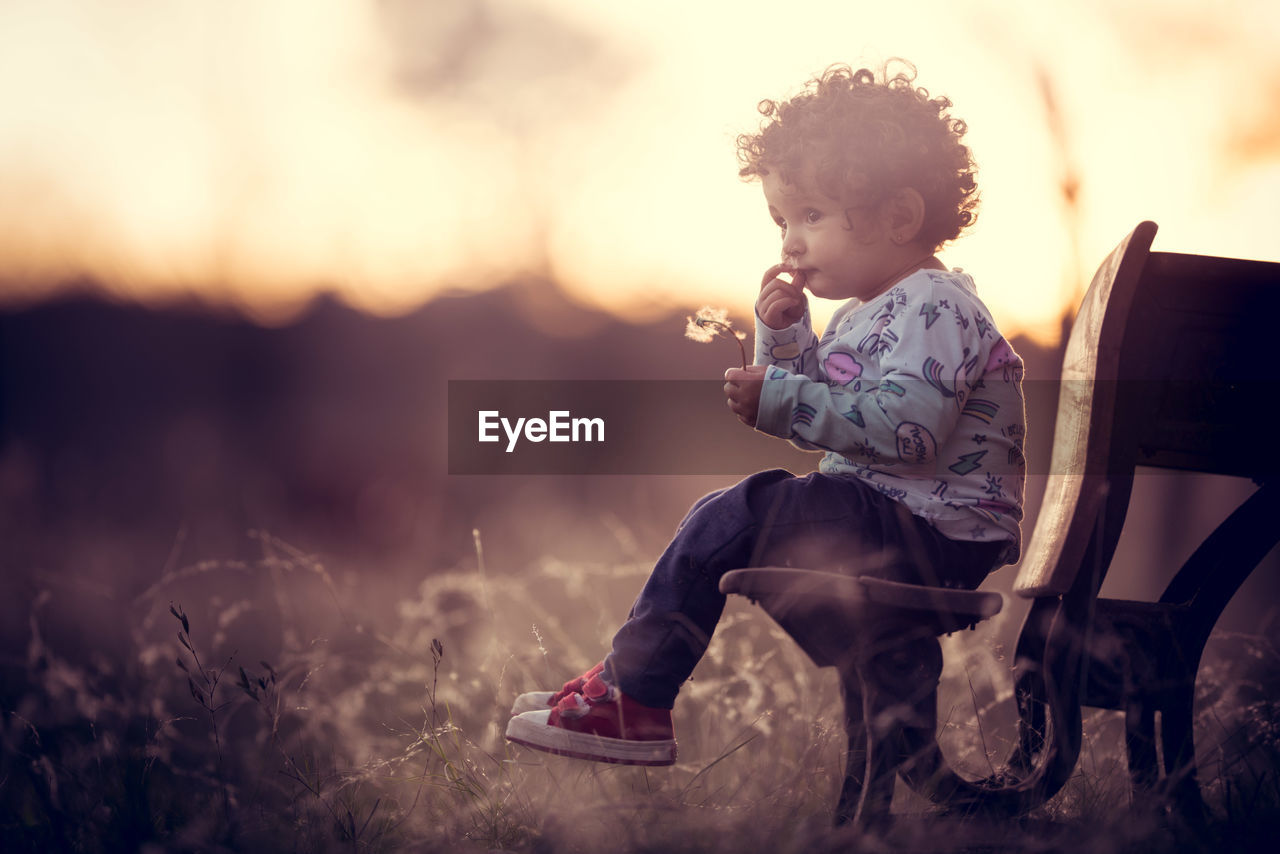 Side view of girl sitting on bench at park during sunset