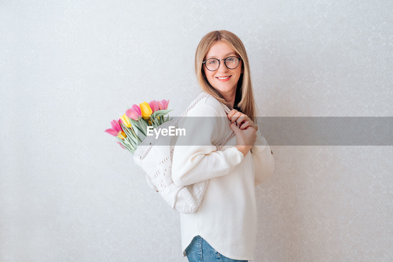 PORTRAIT OF A SMILING YOUNG WOMAN STANDING AGAINST WALL