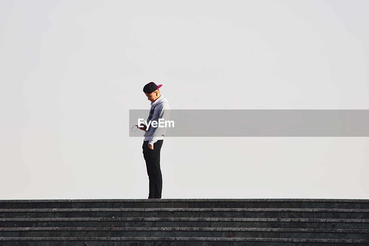 Young man using on mobile phone while standing on steps against clear sky