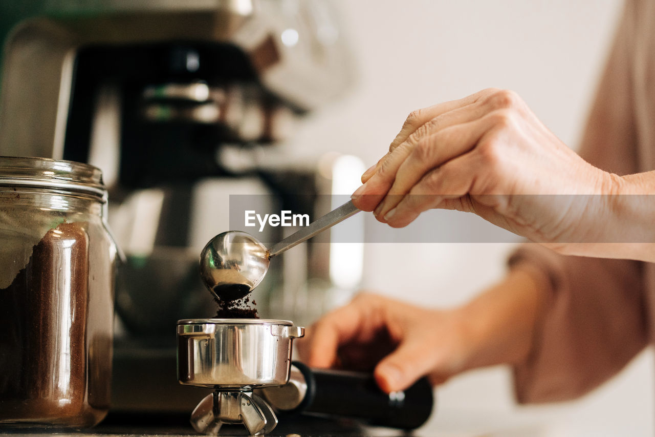 Unrecognizable female with spoon pouring ground coffee into portafilter while standing at kitchen counter with jar of coffee and coffee machine on blurred background