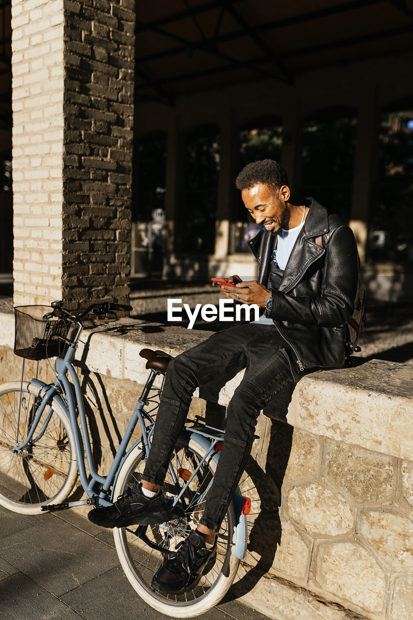 Full body side view of cheerful african american male with backpack sitting with bicycle on paved street near building in city