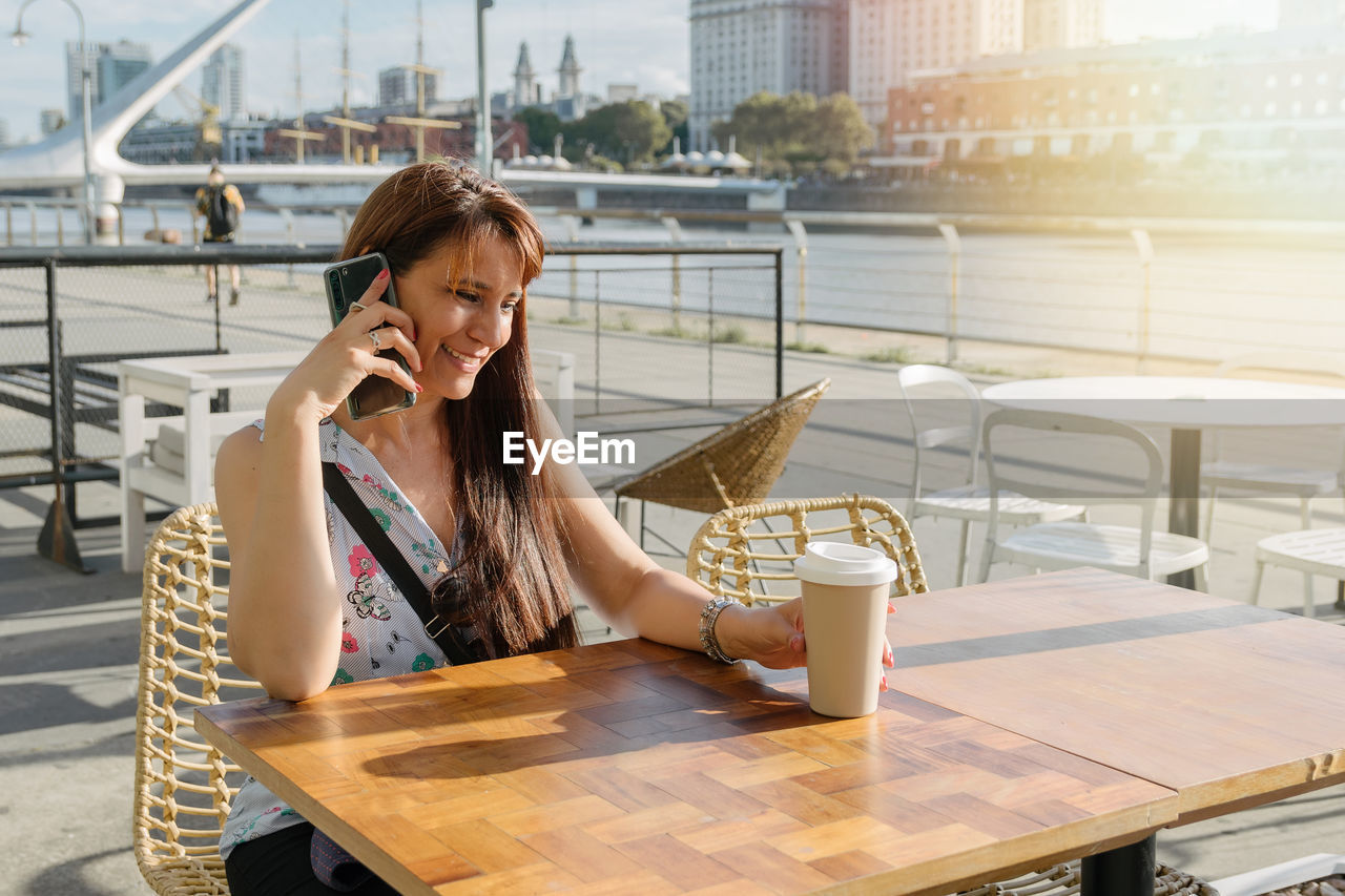 A hispanic woman talking with her phone on cafeteria outside drinking coffee.