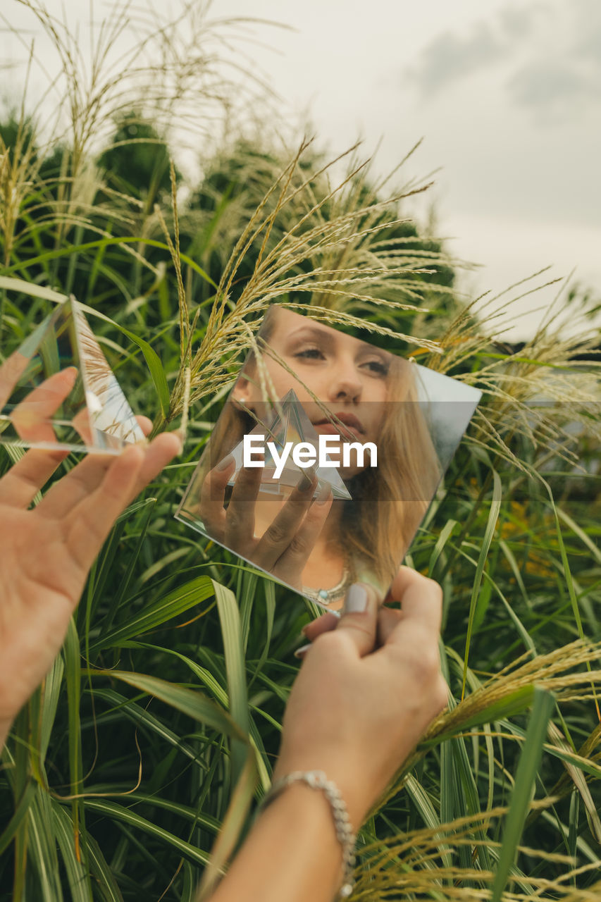 Reflection of woman looking away and holding prism in mirror against plants