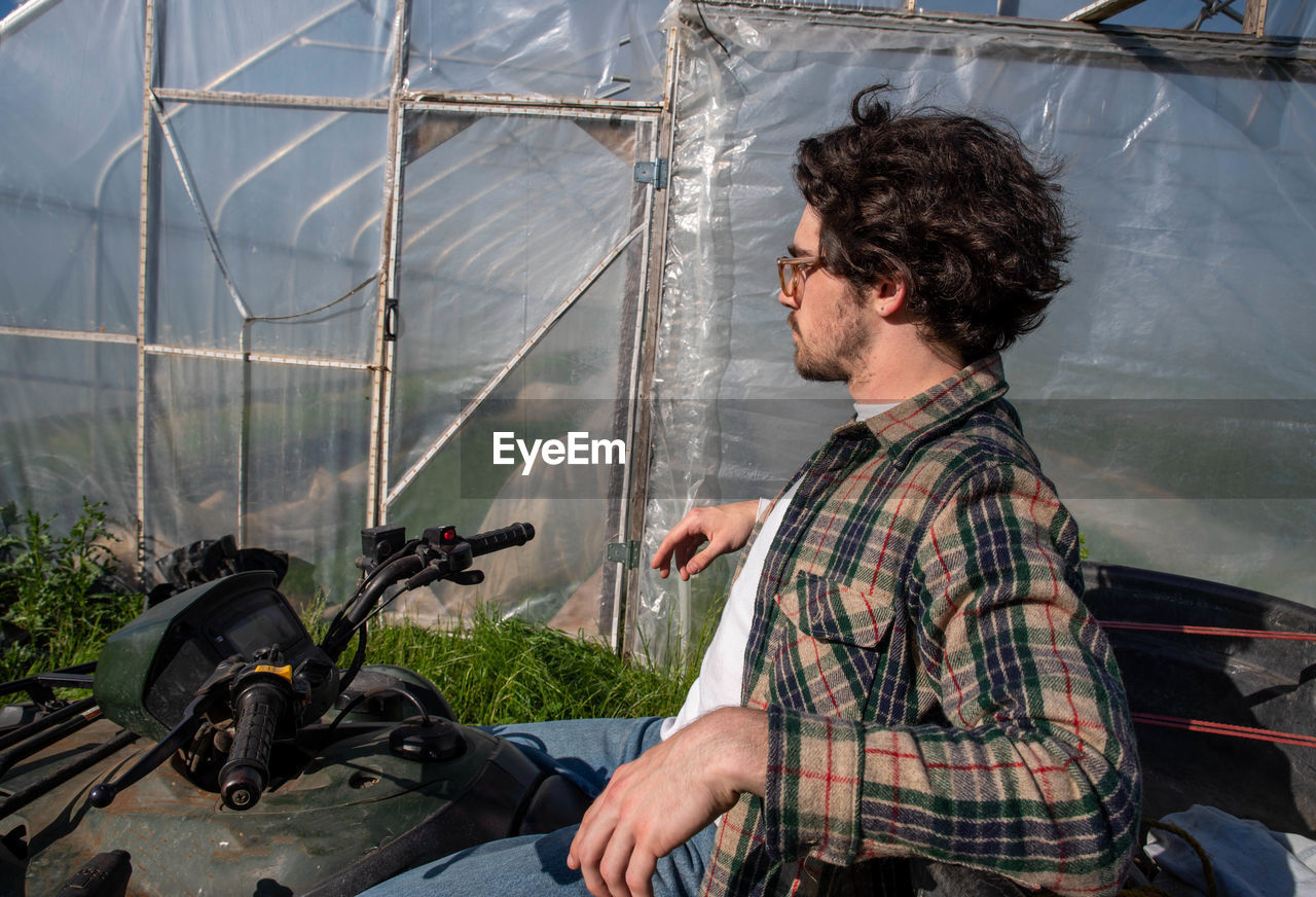 Young farmer in flannel shirt sits on off road vehicle outside greenhouse