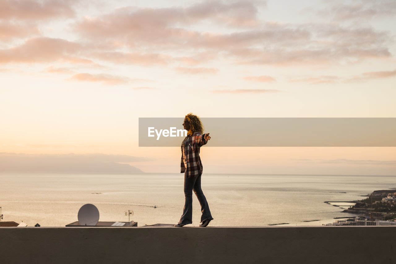 Rear view of woman standing at beach against sky during sunset