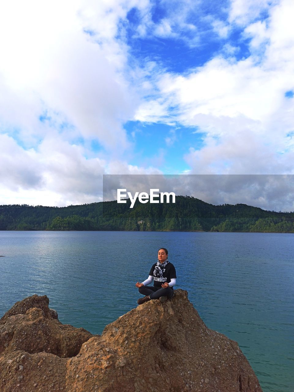 PERSON SITTING ON ROCK BY LAKE AGAINST SKY