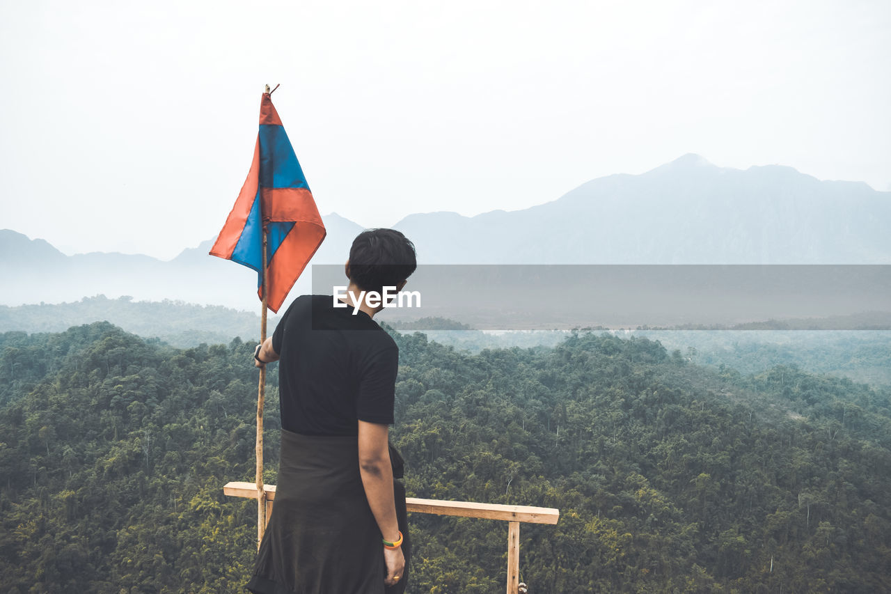 Rear view of man by flag standing on mountain against sky