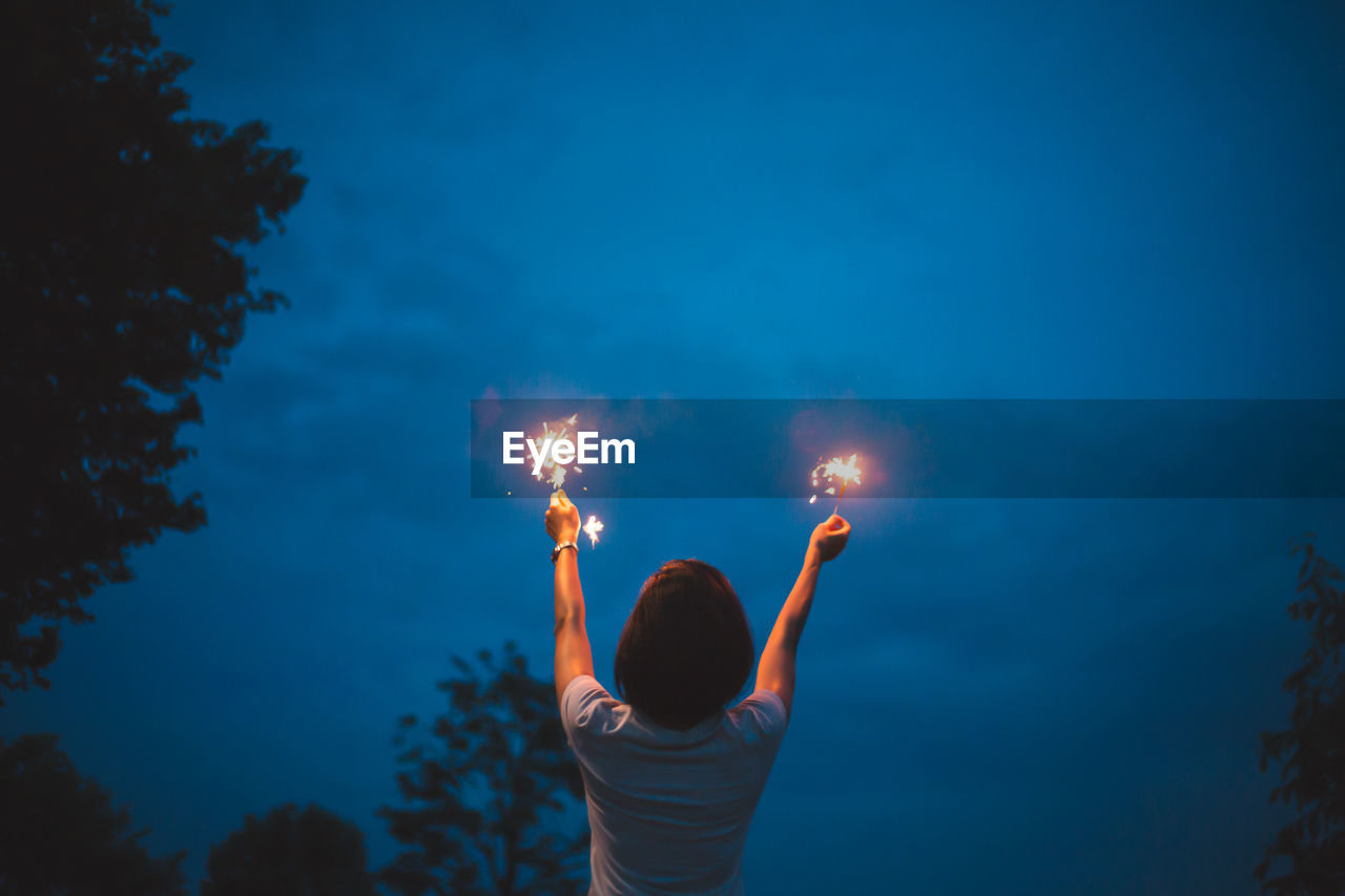 Rear view of woman with arms raised holding illuminated sparklers against sky