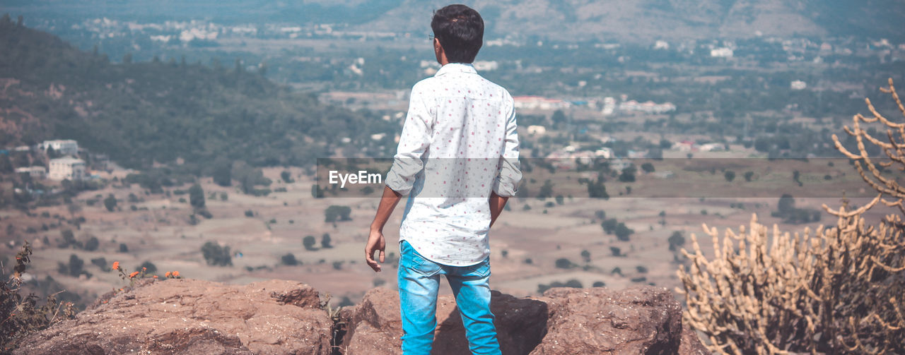 REAR VIEW OF MAN STANDING ON ROCK LOOKING AT VIEW