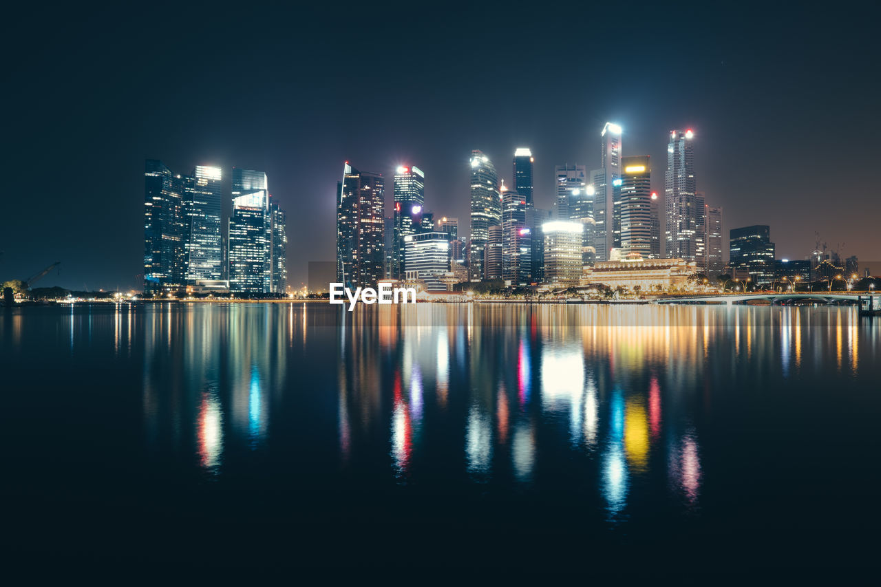 Illuminated buildings by river against sky at night