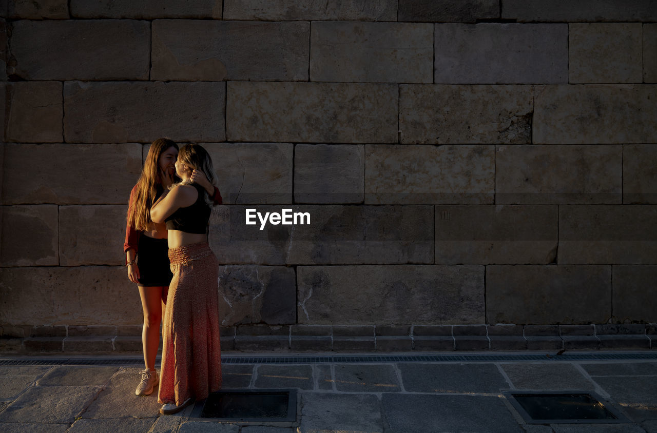 Two girls standing against stone wall during sunset