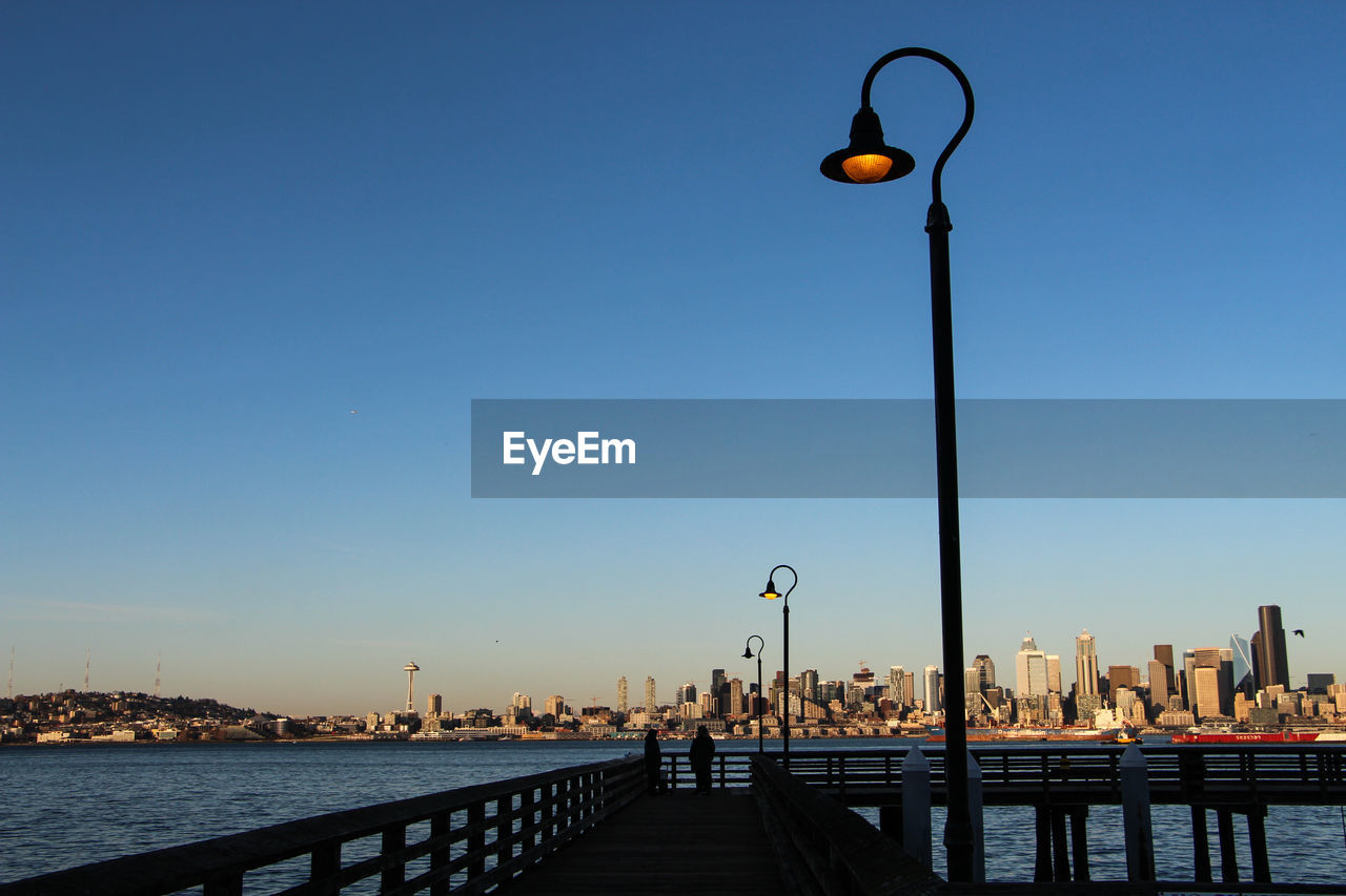 Illuminated street light by city against clear blue sky