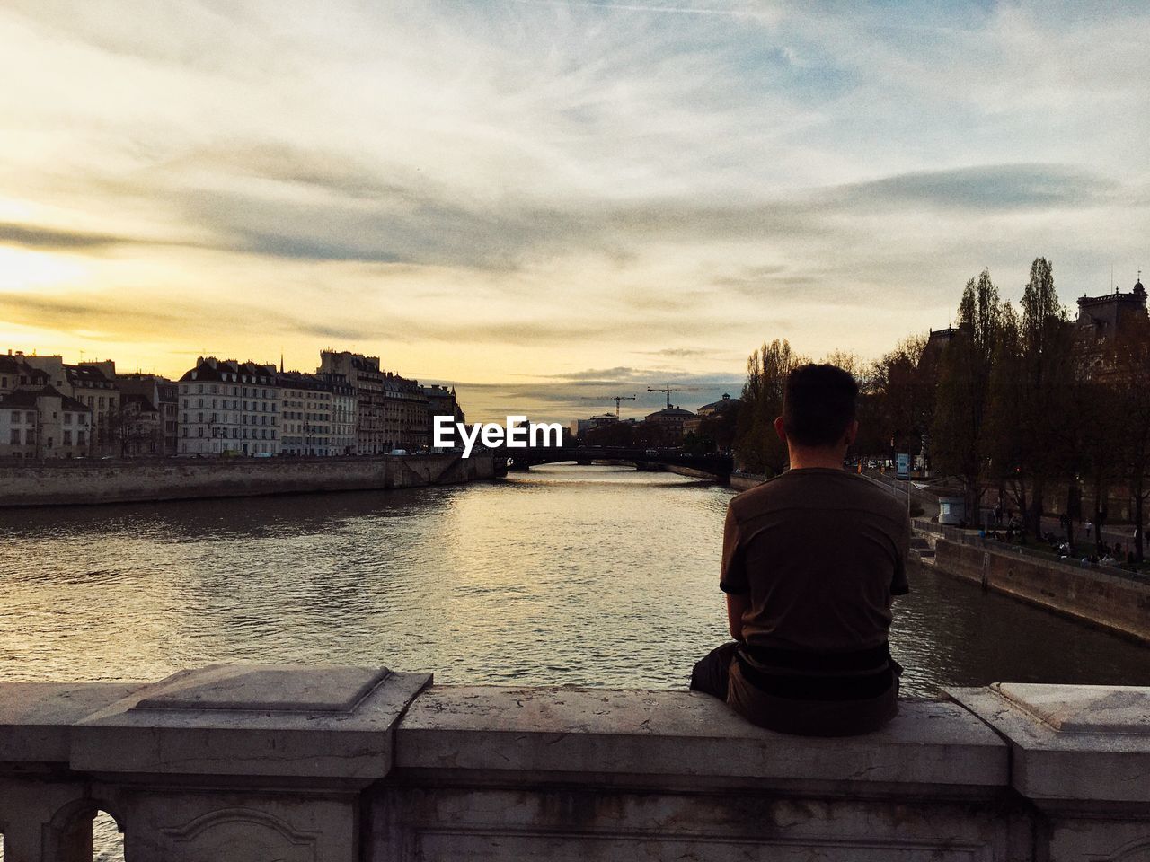 Rear view of man sitting on railing over river against cloudy sky during sunset