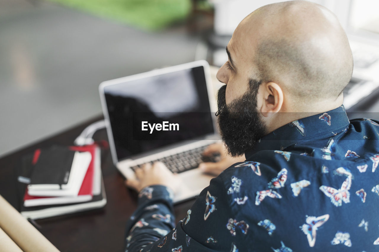 Rear view of male architect looking away while using laptop at table