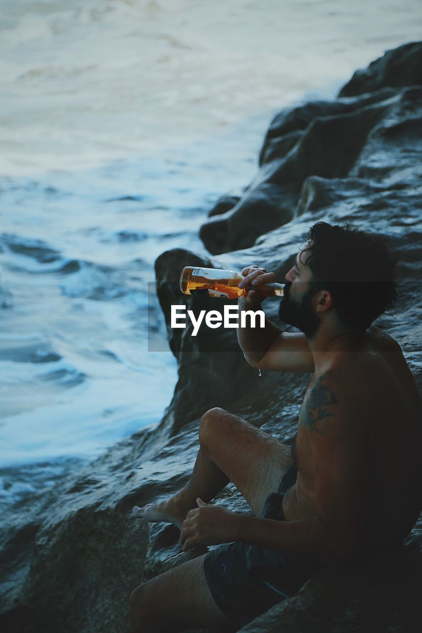 Side view of shirtless mid adult man drinking beer while sitting on rock formation