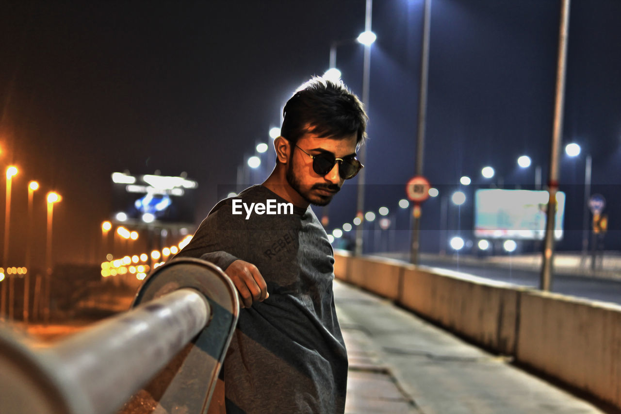 Portrait of young man wearing sunglasses while standing by railing at night