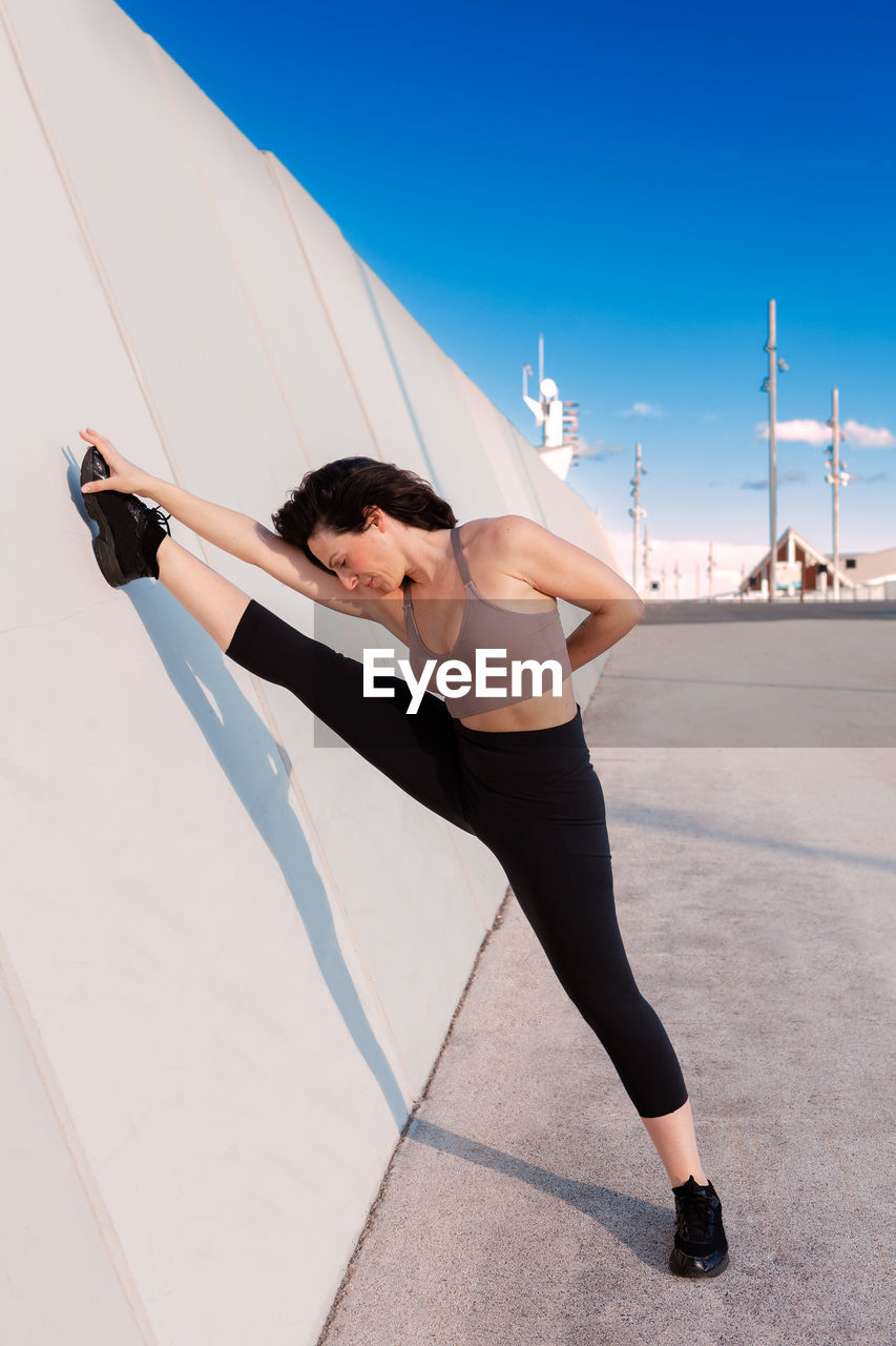 Full body of sporty female in activewear with eyes closed and raised arm doing split and stretching leg during outdoor training against blue sky on summer day