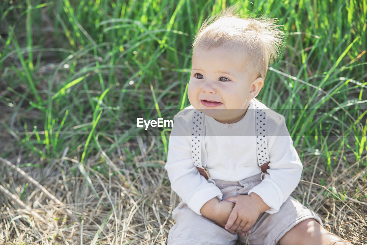 Beautiful blond baby in a suit with suspenders sits in the grass