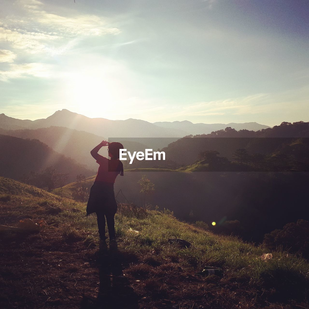 Rear view of woman shielding eyes while standing at mountain against sky during sunset