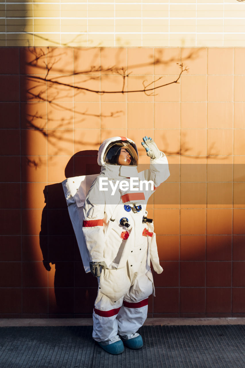 Female astronaut in space suit shielding eyes from sunlight while standing by wall