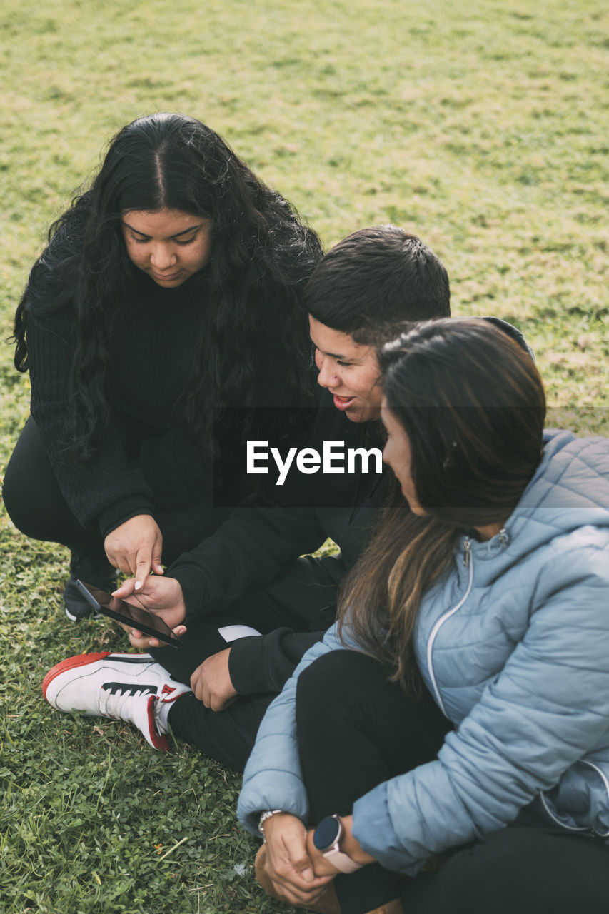 Three people of hispanic-latino ethnicity, sitting on the ground in the park with smart devices