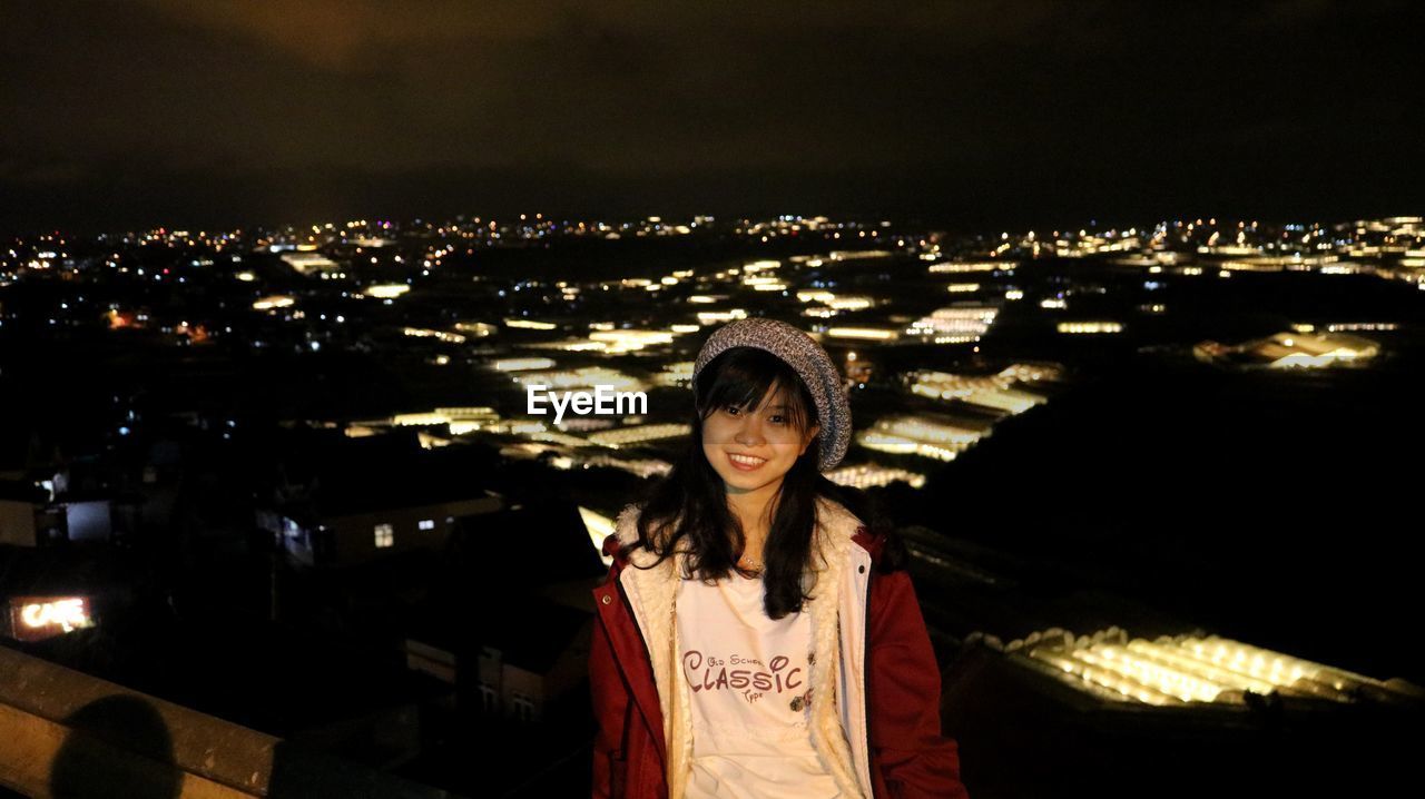 PORTRAIT OF SMILING YOUNG WOMAN STANDING AT NIGHT