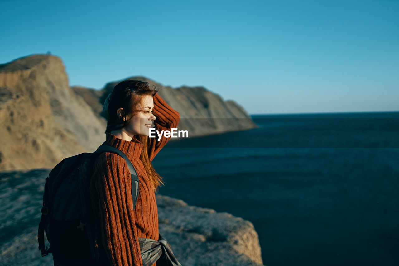 REAR VIEW OF MAN STANDING ON ROCK AGAINST SEA