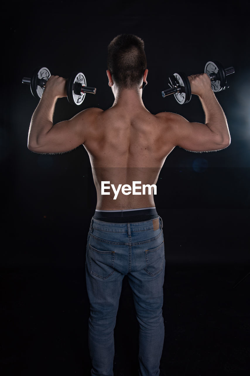 Rear view of shirtless muscular man exercising with dumbbells against black background