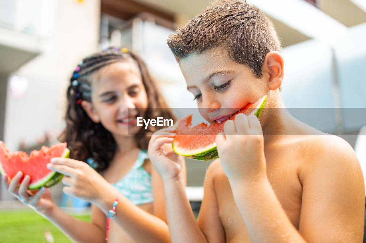 Two funny kids eating watermelon at the the edge of the pool on a summer day