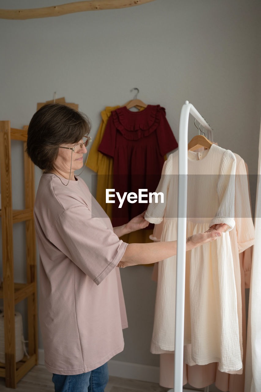 Middle aged woman in casual clothes and glasses examining sleeves of handmade dresses while working in light workshop